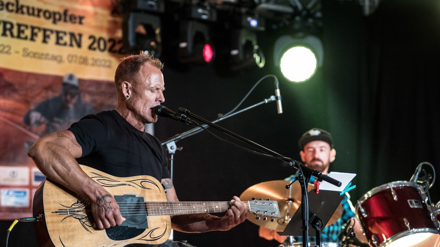 Ein Musiker mit Gitarre und Mikrofon und ein Schlagzeug auf einer Bühne, dahinter Lichttechnik, alles von MSL Eventtechnik.